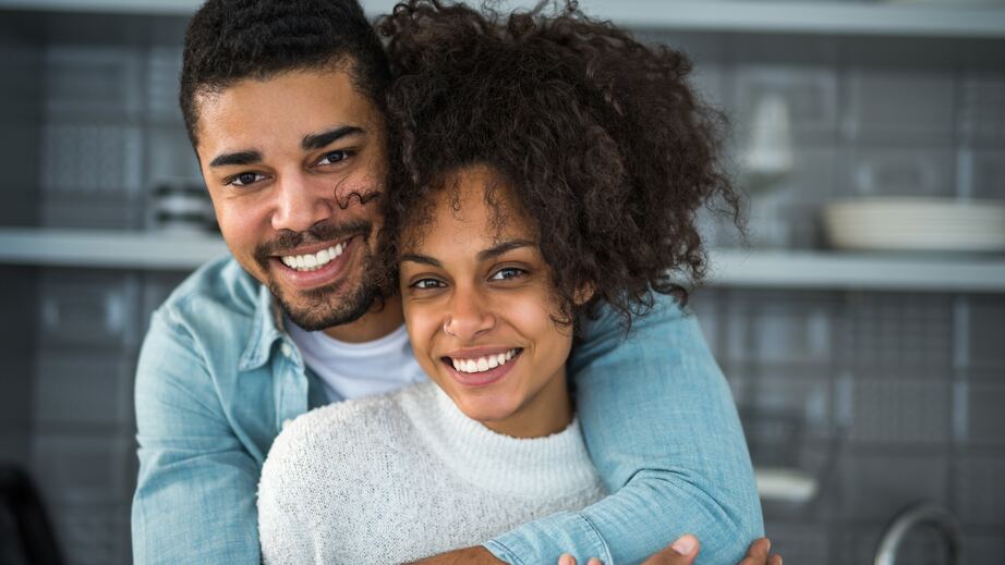 Una pareja joven sonríe a la cámara en su cocina; él la abraza por detrás.